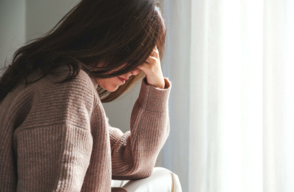 A patient wearing a brown sweater presses their left hand to their forehead, feeling discomfort from a dry eye-induced headache.