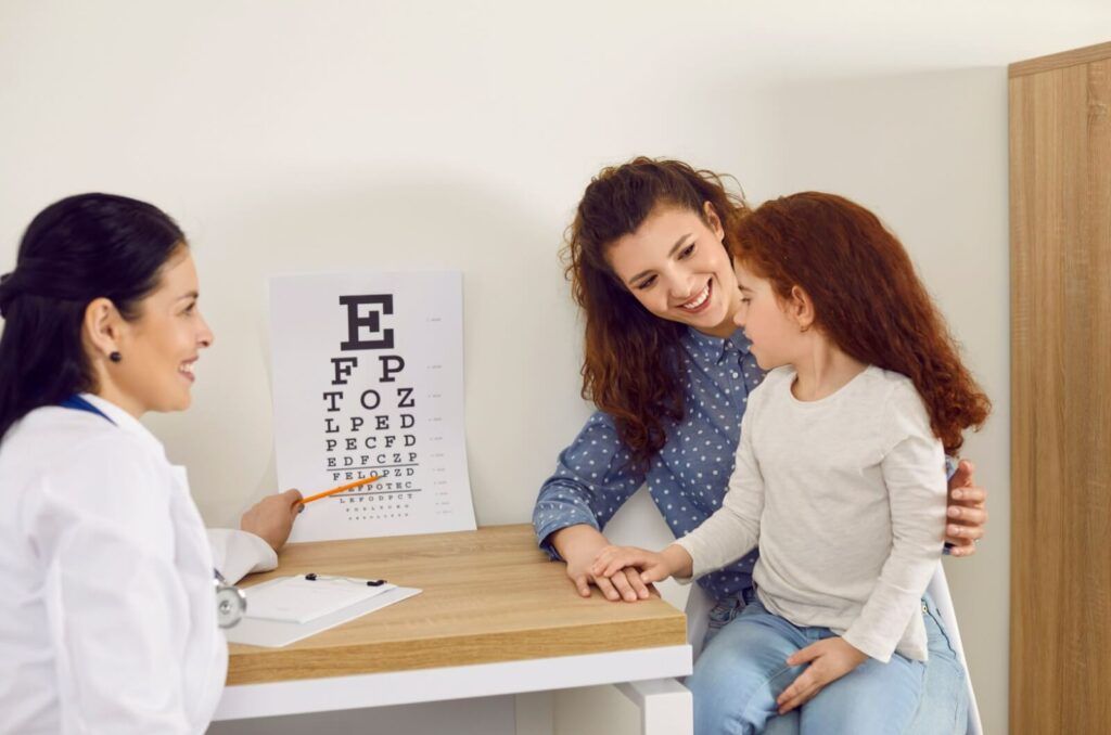 A parent and child attend a child's eye exam; the eye doctor points to a Snellen chart while smiling.