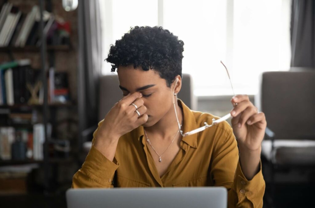 A person working at a laptop removes their glasses and rubs their dry eyes.