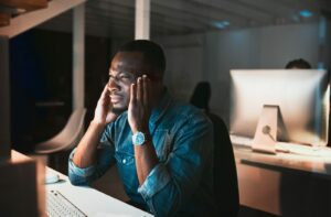A professional rubs their temples to relieve eye strain while staring at a computer in a dimly-lit office environment.