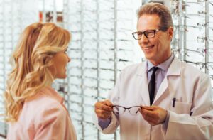 An optometrist holds out a pair of glasses for their client to try on in front of a large display of high-quality eyeglass frames.