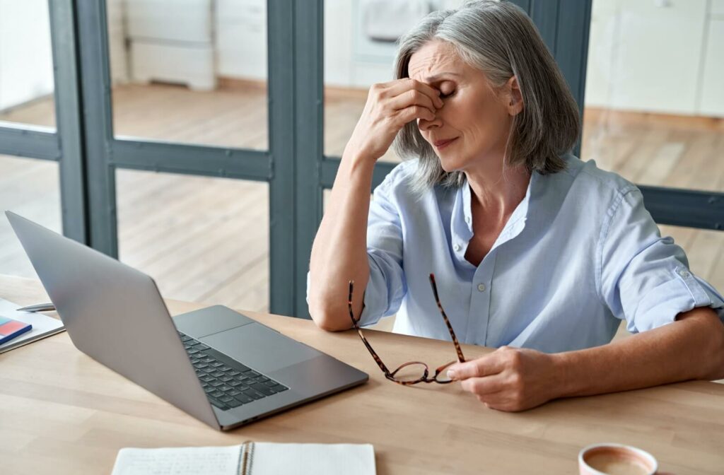 Older woman sitting at a desk with a laptop, holding her glasses and rubbing her eyes from fatigue or eye strain.