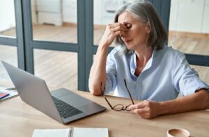Older woman sitting at a desk with a laptop, holding her glasses and rubbing her eyes from fatigue or eye strain.