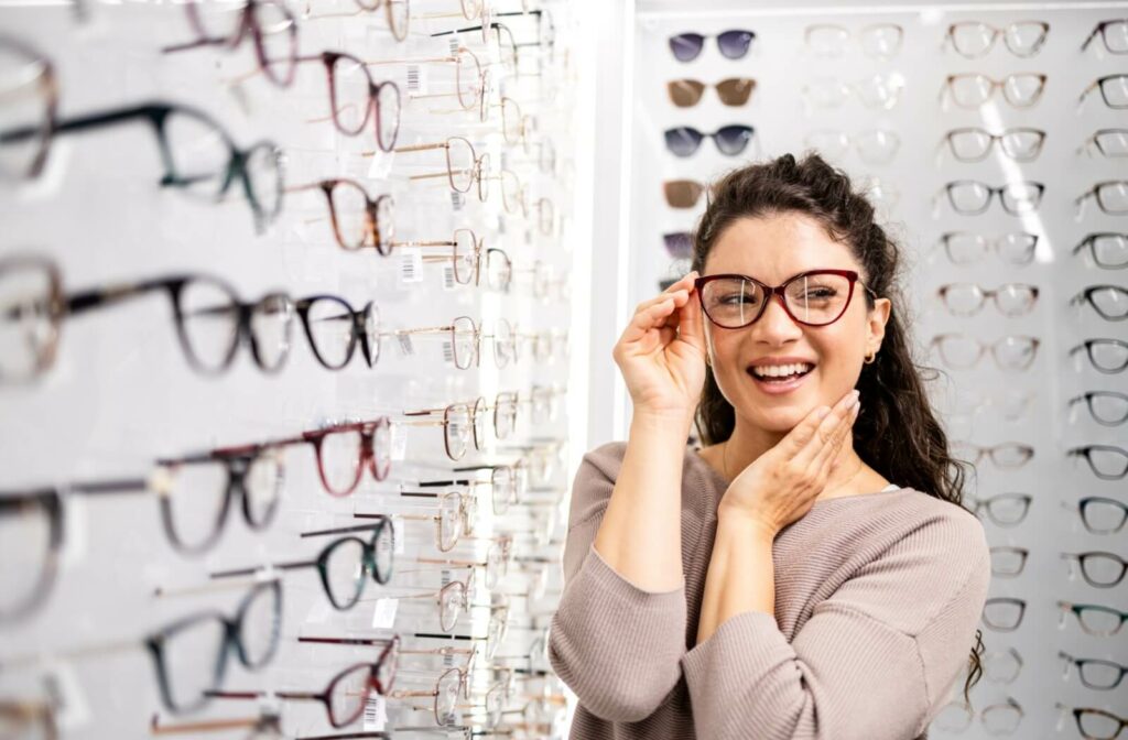 A person with dark, curly hair wearing a lavender top, smiling while trying on dark red cat-eye glasses in front of a wall display of various eyeglass frames.