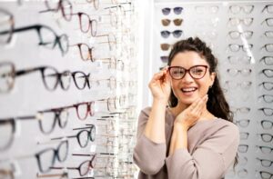 A person with dark, curly hair wearing a lavender top, smiling while trying on dark red cat-eye glasses in front of a wall display of various eyeglass frames.