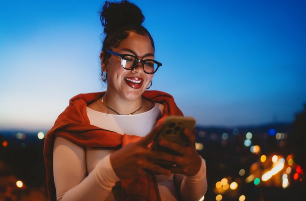 A person with their hair in a bun smiling and using a smartphone at night while wearing stylish black-rimmed eyeglasses.