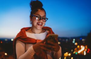 A person with their hair in a bun smiling and using a smartphone at night while wearing stylish black-rimmed eyeglasses.