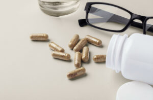 A white bottle of eye health supplements spilled open with several capsules next to a pair of black eyeglasses.