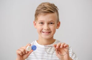 A smiling young boy holding a contact lens case in one hand and a pair of eyeglasses in the other.