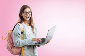 A smiling young girl wearing black-framed eyeglasses and a backpack while holding a laptop against a pink background.