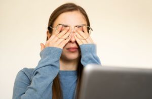 A young woman wearing clear-framed glasses rubbing her eyes in front of a laptop, illustrating symptoms of dry eye syndrome and digital eye strain.