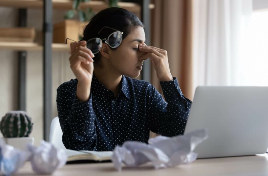 A woman sitting at a desk with a laptop, holding her glasses and pinching the bridge of her nose to relieve dry eye discomfort and fatigue.
