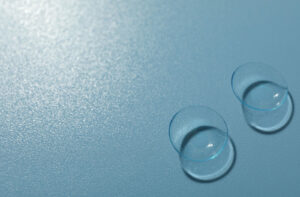 A close-up view of two clear contact lenses resting on a textured light blue surface, illustrating considerations for contact lens wear during allergy flare-ups.