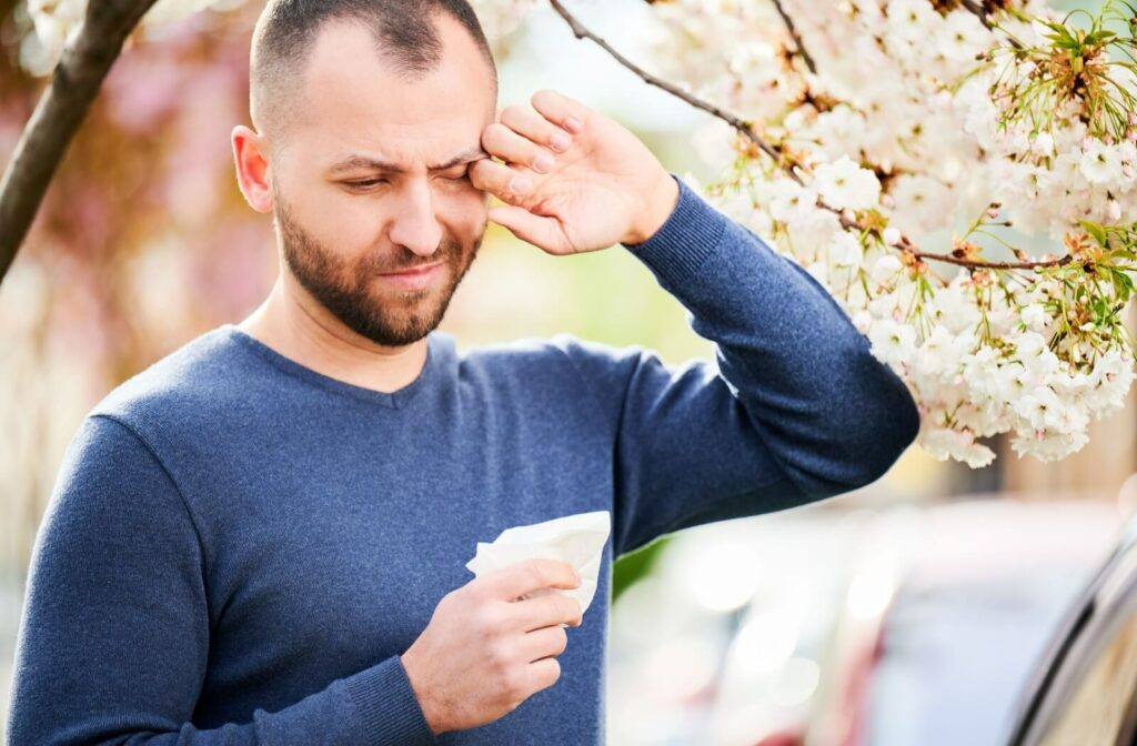 A man standing outdoors rubbing his itchy, swollen eye next to blooming white spring blossoms while holding a tissue, illustrating seasonal allergic conjunctivitis symptoms.